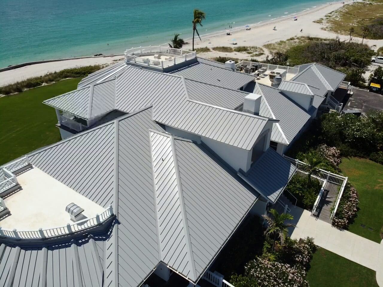 Close aerial angle of a standing-seam metal roof system on a beachfront residence, showing multiple hips, valleys, and roof-mounted equipment