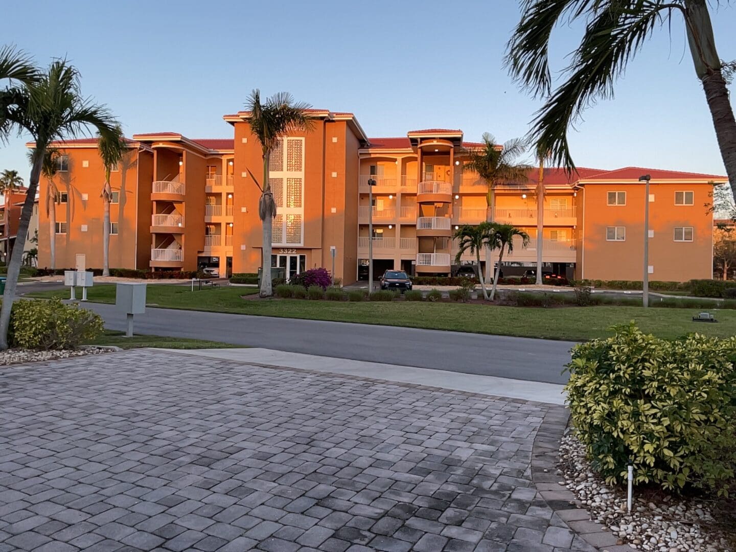 Street-level view of a multi-story orange condominium building with tile roofs, exterior stairwells, and a paver driveway lined with palm trees